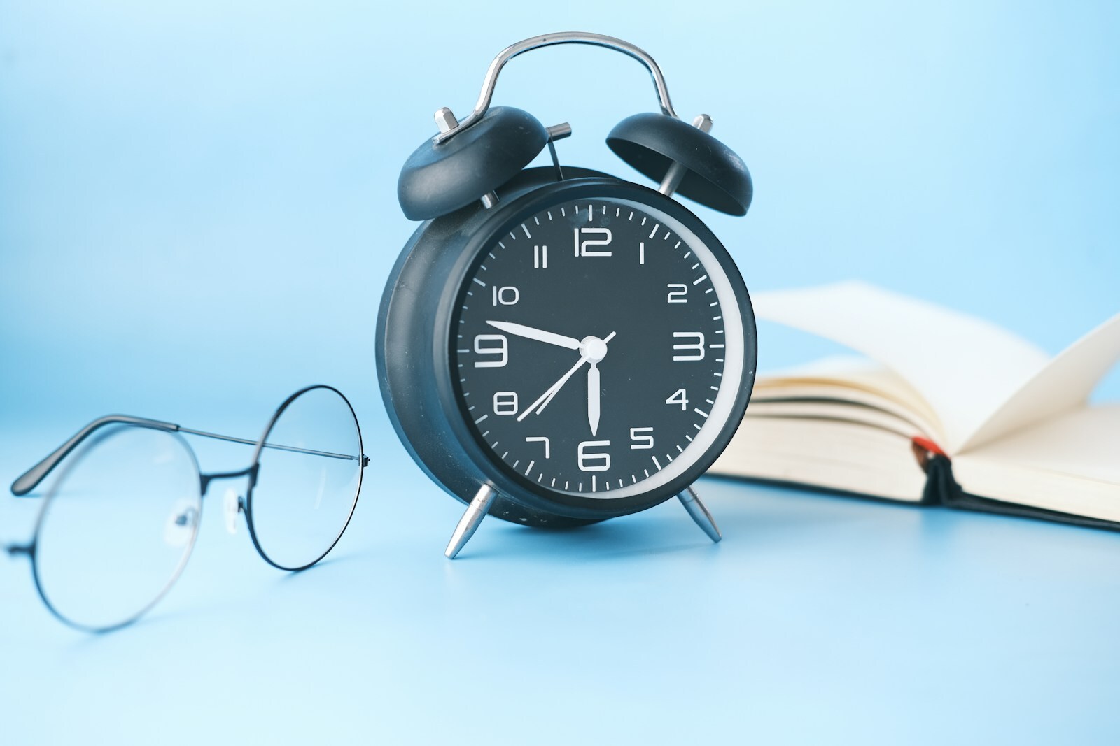 An analogue alarm clock on a surface with a pair of spectacles and and open book.